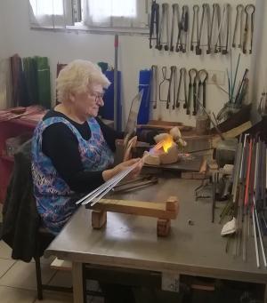 Photo d’un maitre verrier à Murano, une femme âgée fabriquant des perles de verre artisanales pour un chapelet, représentant l’art ancestral du verre vénitien.
